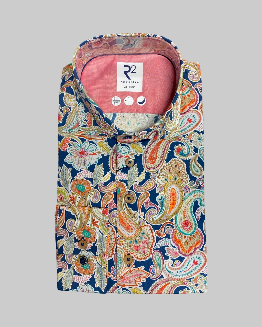 A folded long-sleeve button-up shirt by R2 Lifestyle, featuring a navy blue base with a vibrant multicolor paisley pattern in orange, green, beige, and white. The inside collar reveals a pink lining and a white brand label.